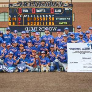 The Pomona-Pitzer men’s baseball team gather on the field in front of the score board with their NCAA trophy.